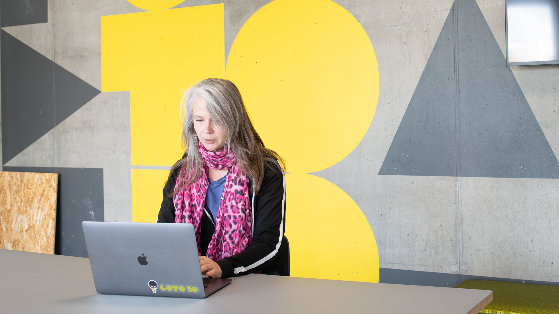 Kvinna sitter vid bord och skriver på sin laptop och sitter framför Goto 10-målning på väggen