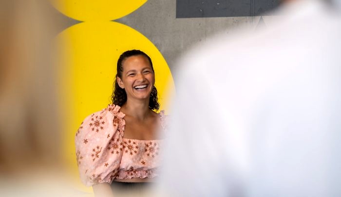 Skrattande kvinna står framför målad tegelvägg i gult och tittar in i kamera m