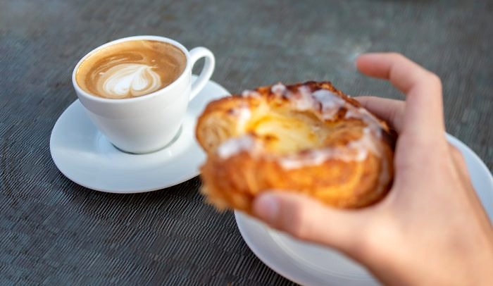 en kaffekopp med ett fat och en hand som håller i ett Wienerbröd